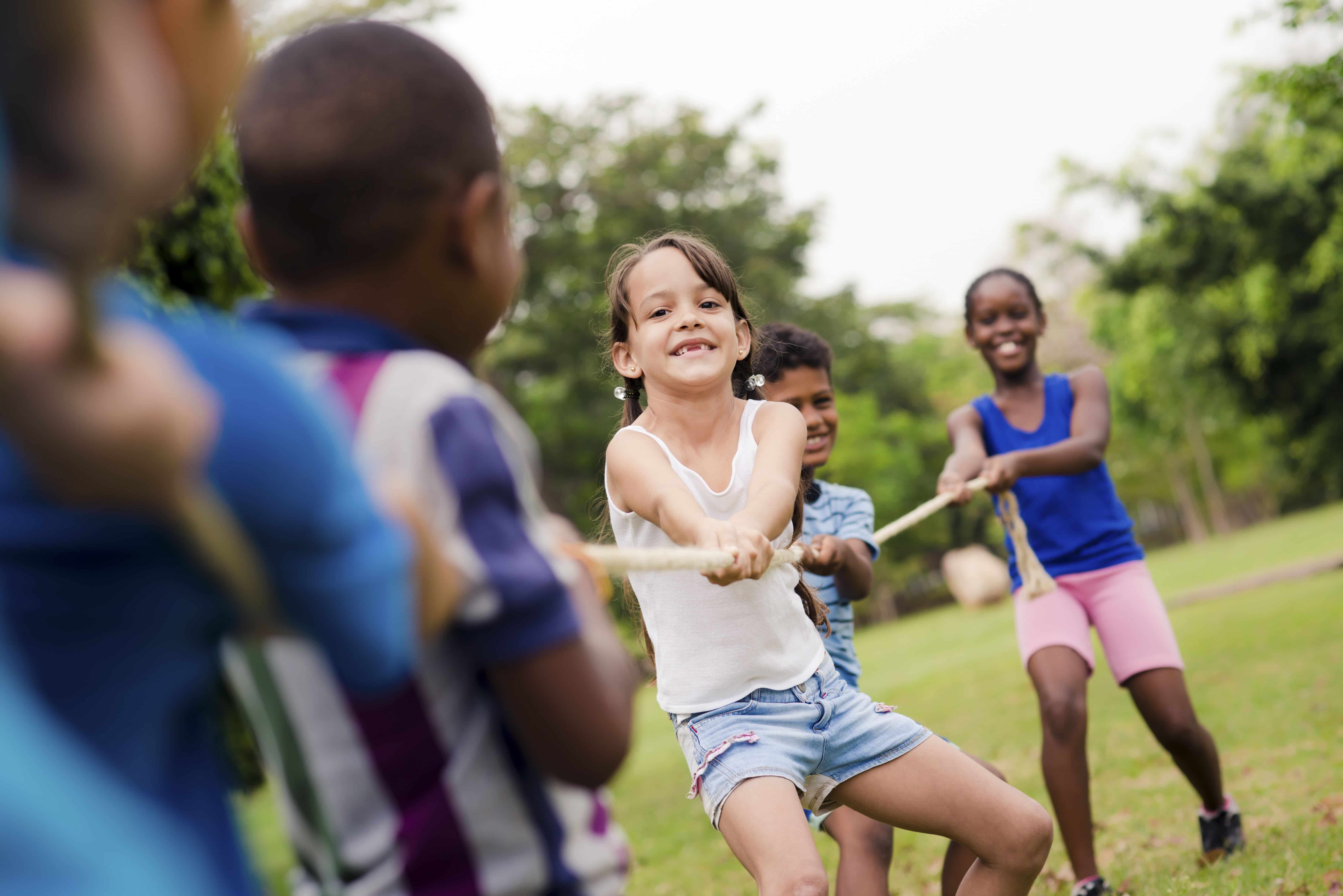 Young kids at summer camp playing tug of war. There are 2 girls and a boy on the opposing team smiling as they play.